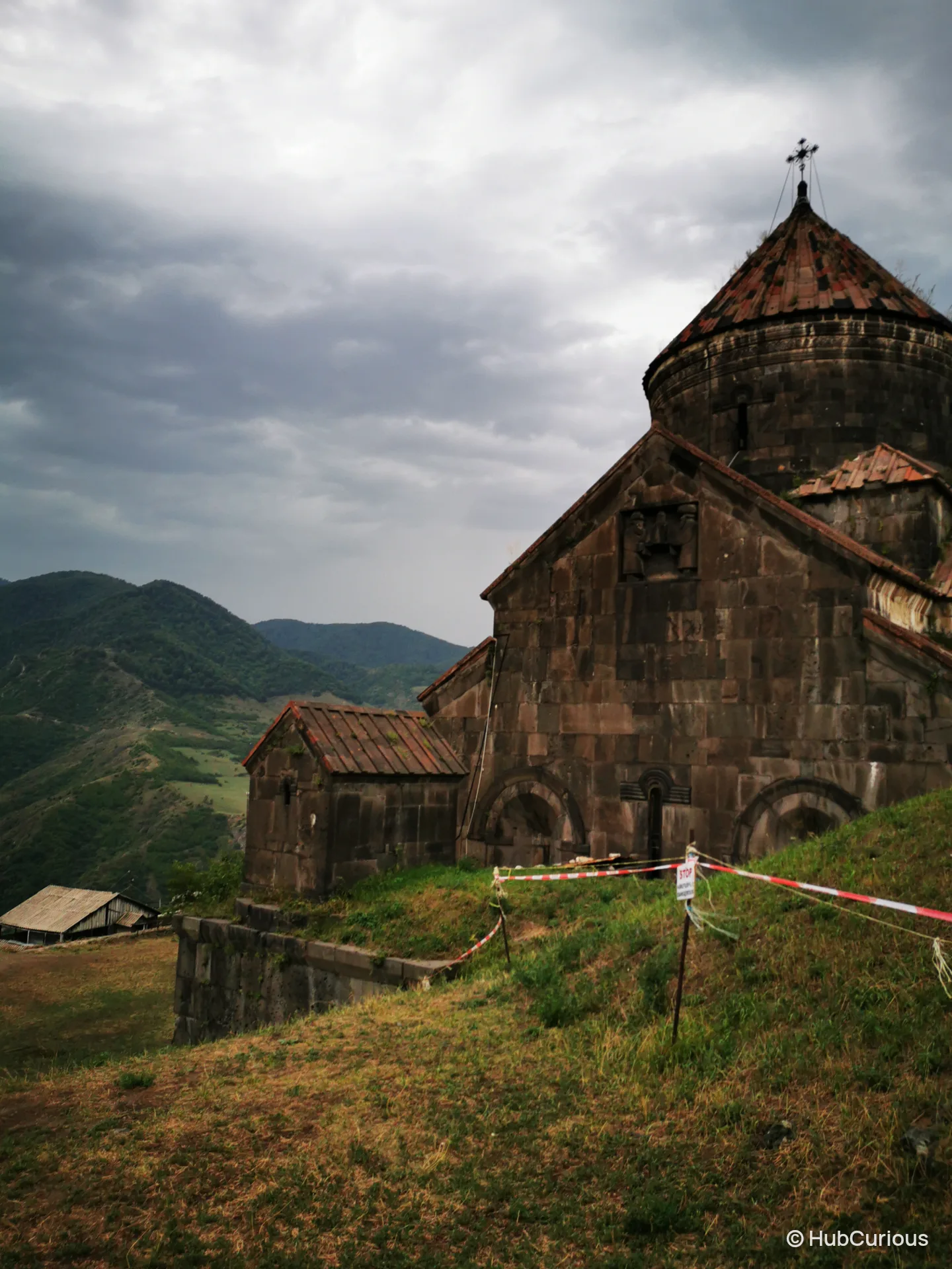 Haghpat Monastery under heavy grey cloud, its rust-red tiled roofs above a grass slope closed off with tape