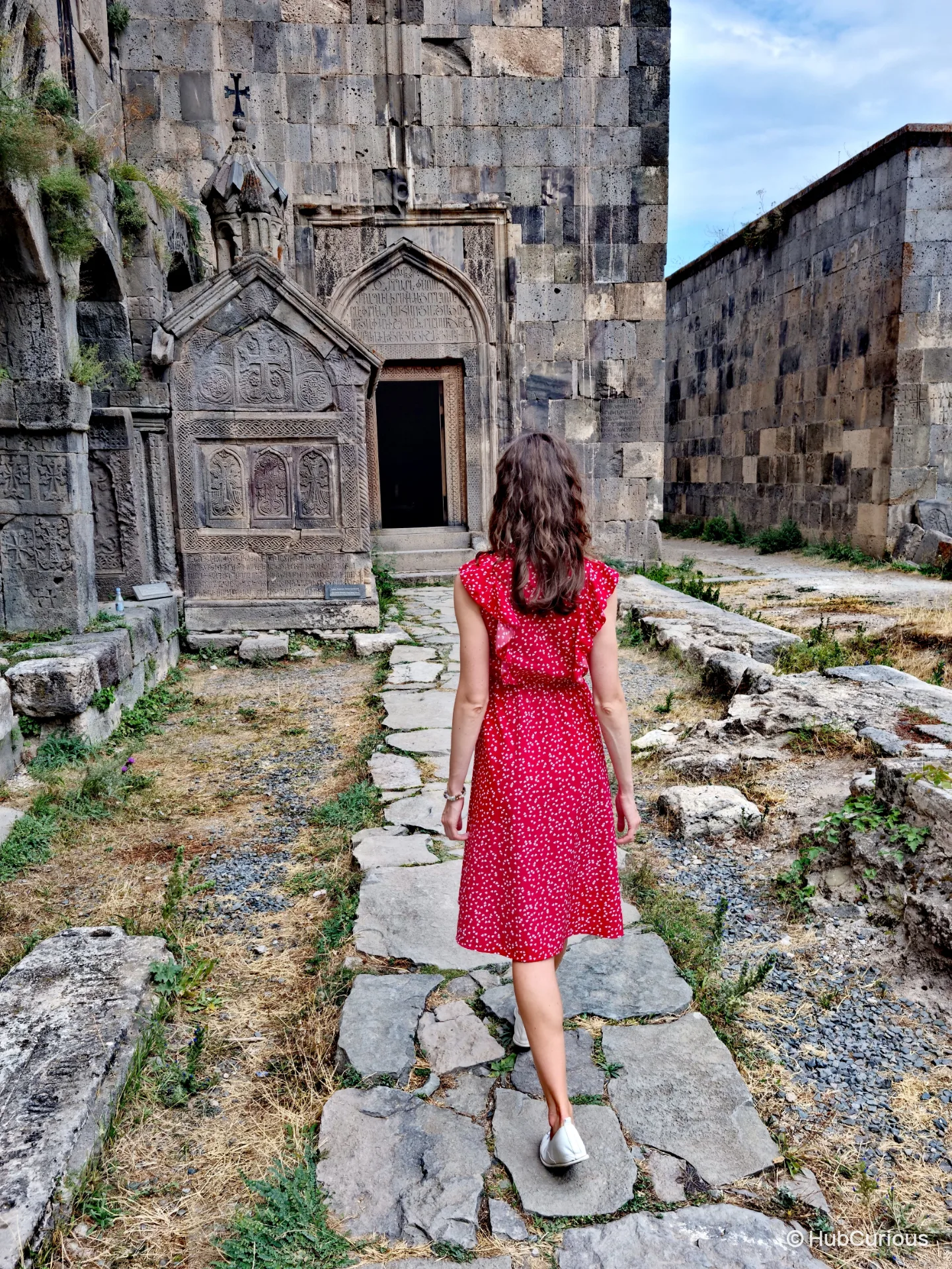 Woman in a red polka-dot dress walking a stone path towards the carved doorway of Tatev Monastery, khachkars along the wall