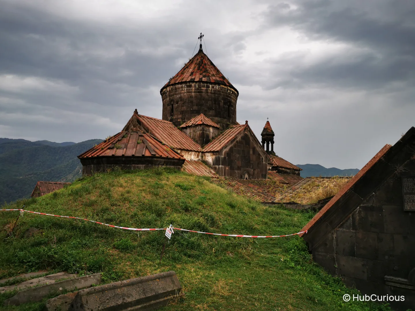 The roofs and drum of Haghpat Monastery seen across a grassy mound, with red and white tape strung along the slope