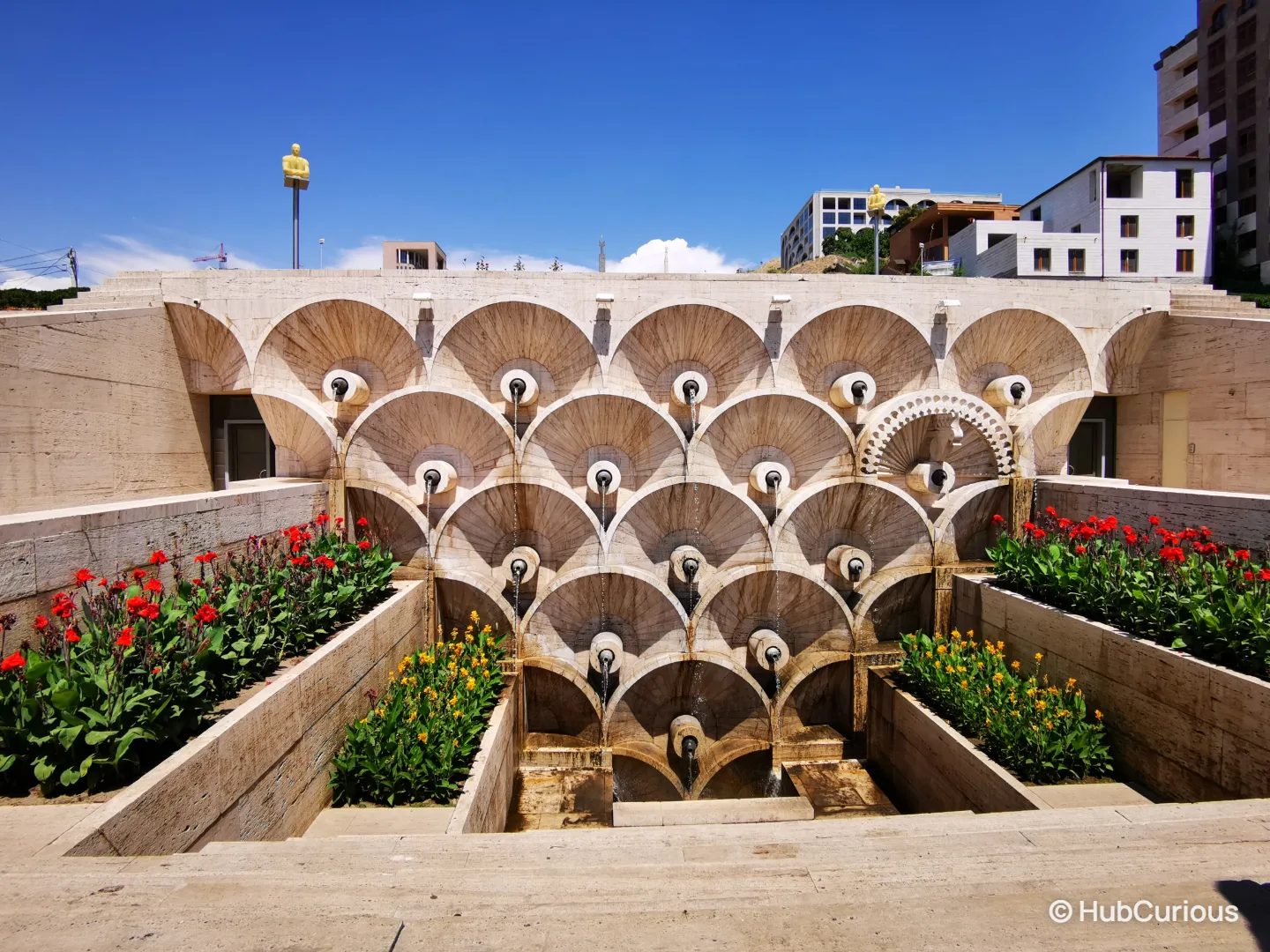 Fountain at the foot of the Cascade in Yerevan, with fan-shaped stone basins and red and yellow flower beds under a blue sky