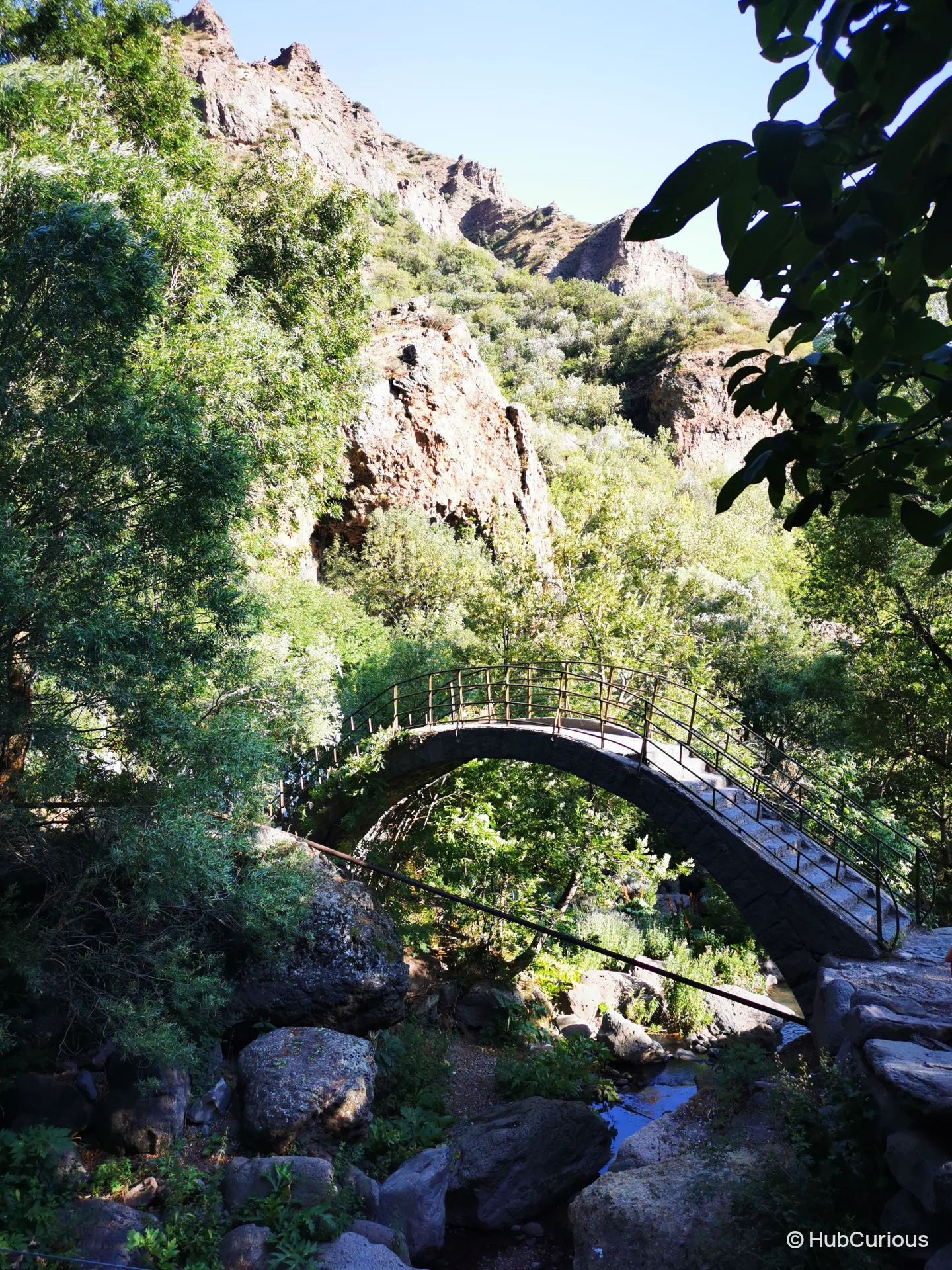 Arched stone footbridge with a metal railing crossing the stream below the cliffs near Geghard