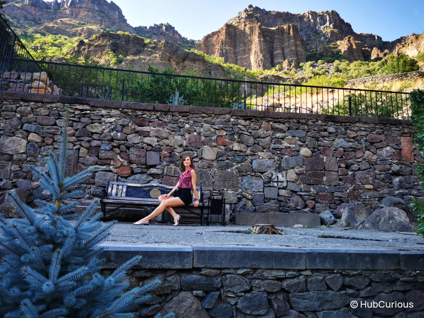 Woman sitting on a bench on the stone terrace at Geghard, with basalt cliffs rising behind the wall